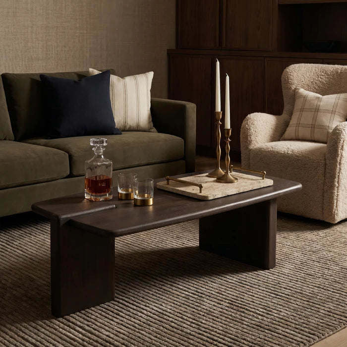 A dark wood coffee table in a styled living room setting, decorated with a glass decanter, two glasses, and a brass candlestick holder on a tray.
