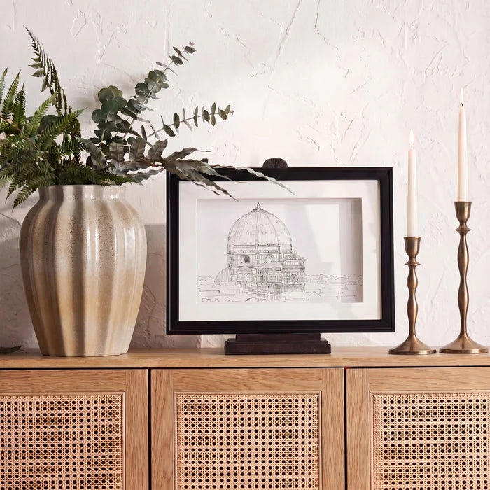 A black-framed art print featuring a detailed sketch of a domed building, styled on a wooden cabinet next to a large beige vase with greenery and two brass candlesticks against a textured white wall.