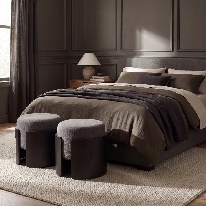A styled bedroom scene with a bed dressed in taupe and dark gray bedding, dark wood paneling on the wall, a bedside lamp, and two matching dark ottomans at the foot of the bed.