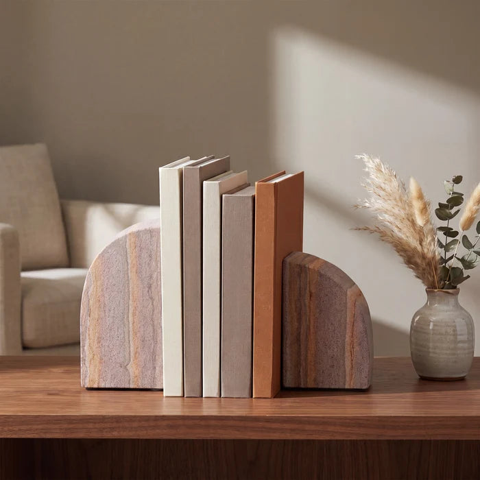Two natural stone bookends holding a set of neutral-colored books on a wooden table, with a blurred living room background and a small vase of dried grass.