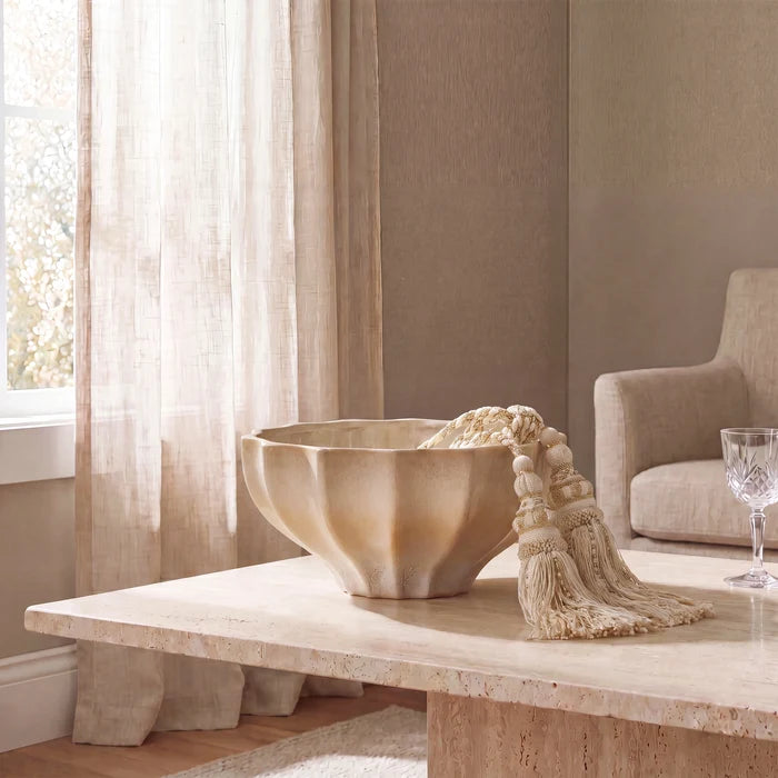 A large scalloped beige stone bowl with a matching tassel runner sits on a light stone table in a softly lit room with sheer curtains, a neutral chair, and a wine glass.