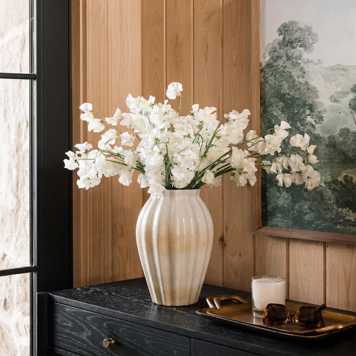 A ribbed ceramic vase with a warm cream and beige gradient, filled with white artificial flowers, displayed on a dark wood console table in a styled interior setting.