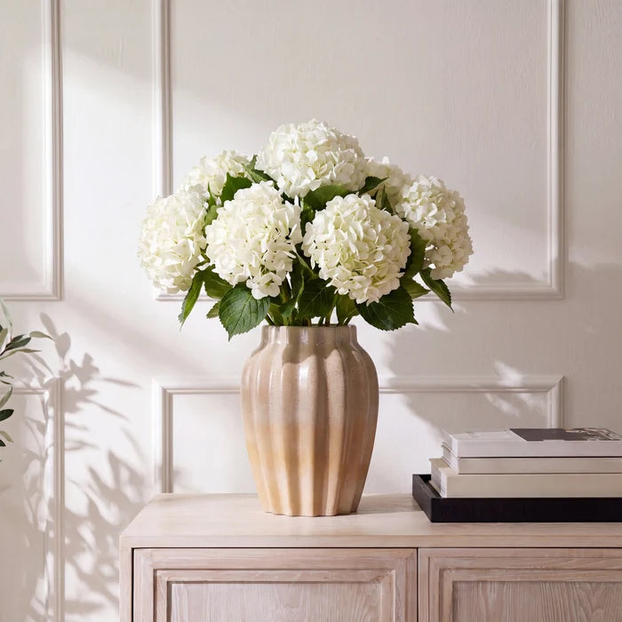 White hydrangea flowers arranged in a ribbed beige ceramic vase, placed on a light wood cabinet against a white paneled wall.