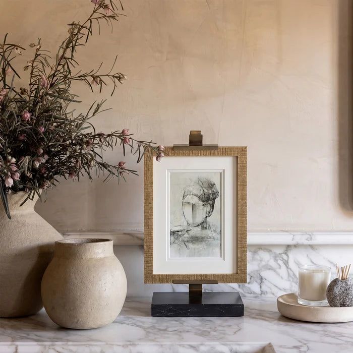 A framed black and white sketch print on a small easel stand, styled on a marble surface with ceramic vases and candles in a warm neutral interior setting.