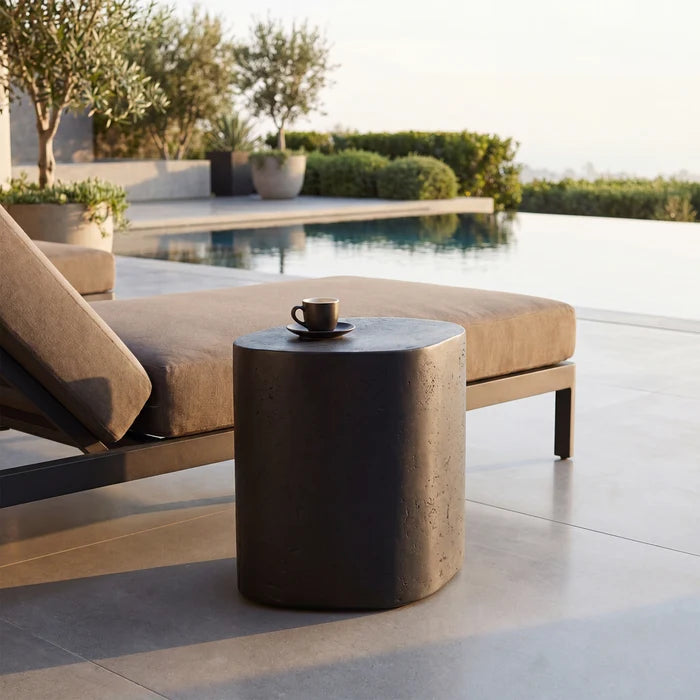 A dark gray cylindrical outdoor side table with a small coffee cup on top, positioned next to a beige cushioned lounge chair on a light stone patio overlooking a swimming pool.