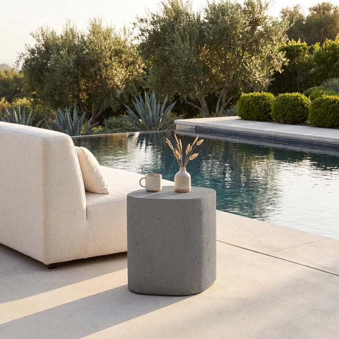 A modern outdoor patio scene with a light beige upholstered lounge chair beside a cylindrical concrete side table holding a small vase and mug, set next to a swimming pool with trees and shrubs in the background.