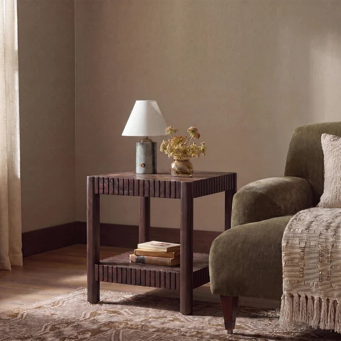 A dark brown wooden end table with a slatted front and lower shelf, styled in a cozy room setting next to an olive green armchair. On the table sits a table lamp with a white shade and a small vase of dried flowers.