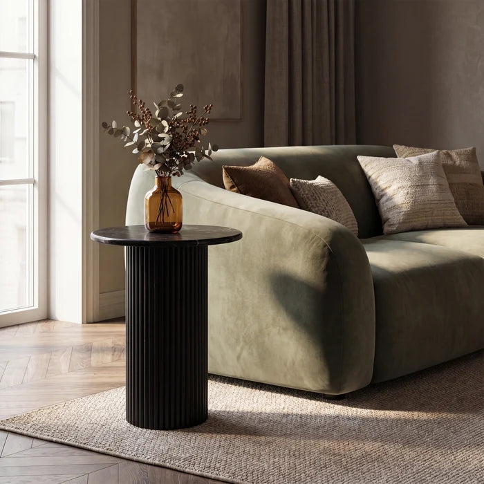 A modern living room scene featuring a ribbed black cylindrical side table next to a green velvet sofa with decorative pillows, styled with an amber glass vase containing dried botanicals.