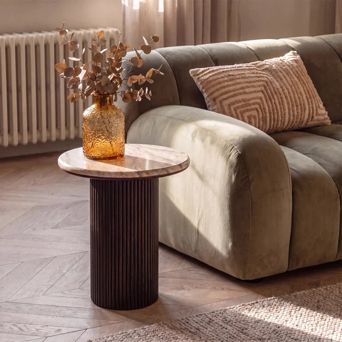 A modern round side table with a fluted dark brown column base and a light marble top, styled in a living room next to a green sofa with a patterned pillow and a glass vase on top.
