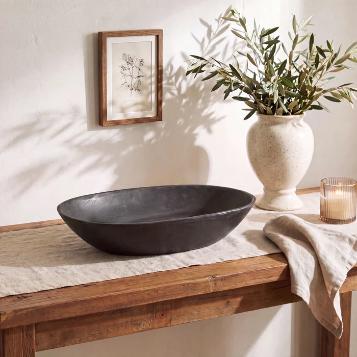 A dark charcoal gray oval stone vessel sink displayed on a rustic wooden console table in a styled bathroom setting. The scene includes a textured cream ceramic vase with olive branches, a lit candle in a glass holder, a beige linen runner, and a small framed botanical print on the wall.