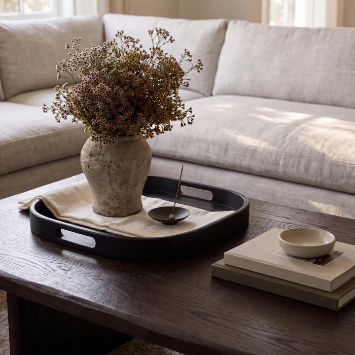 A black rectangular serving tray with cutout handles sits on a dark wood coffee table in a living room setting. The tray holds a beige fabric liner, a textured ceramic vase filled with dried flowers, a small dark bowl, and a candle.