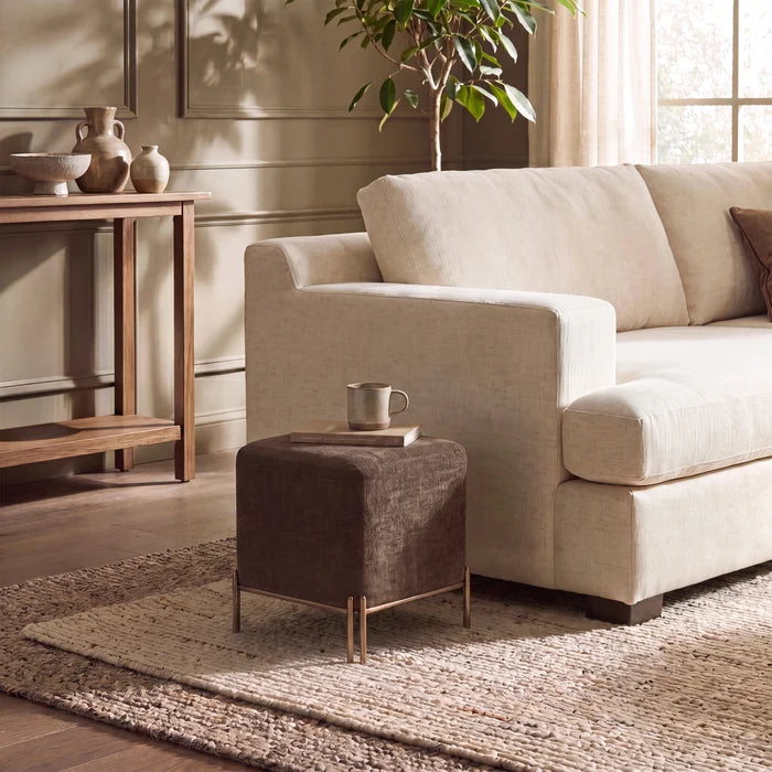 A beige upholstered loveseat sofa in a styled living room, shown next to a small dark brown cube ottoman with gold-toned legs on a woven rug.