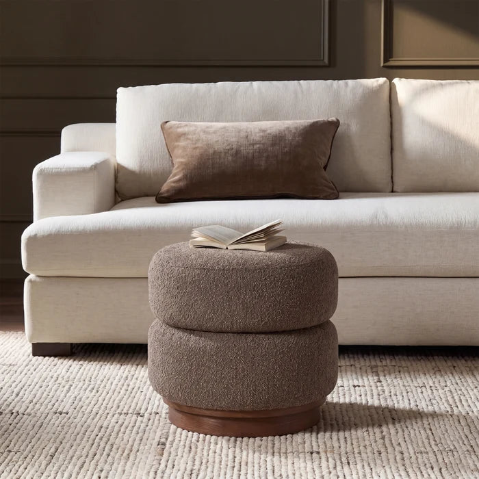 A two-tiered round ottoman in textured taupe bouclé fabric with a dark wood base, styled in a living room setting in front of a cream sofa with a brown accent pillow.