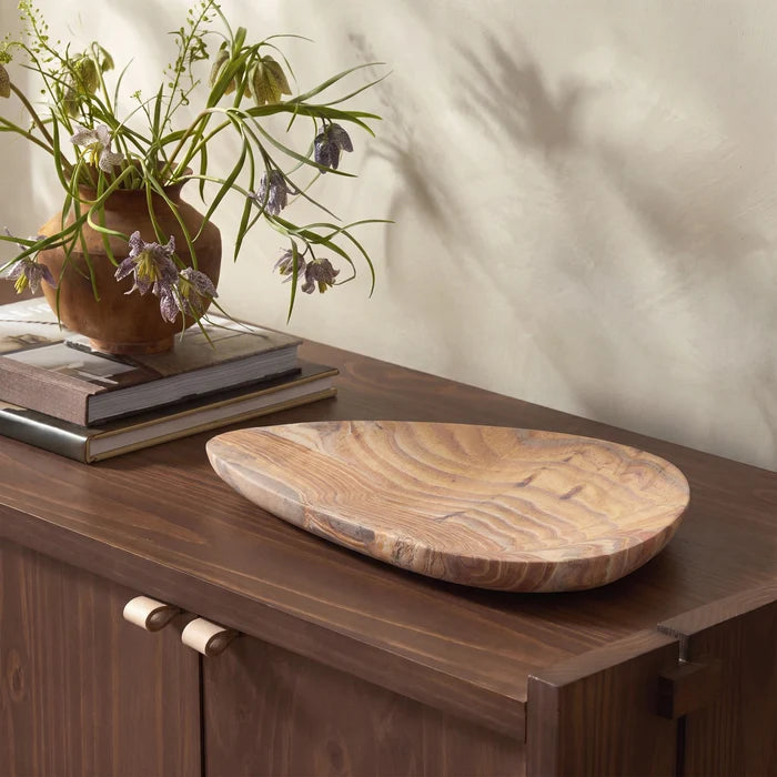 An organic-shaped wooden serving tray with a rich wood grain pattern, styled on a dark wood sideboard next to a ceramic vase with dried flowers.